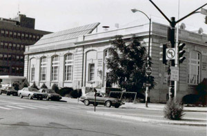 A black and white photo of the light-bricked building with large arched windows and solar panels on the top of the left portion of the building, in the background is a building on the left.