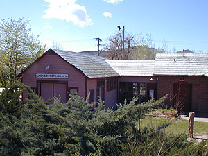 A photo of the building with gabled roof and extension. In the foreground is a large bush on the bottom left corner.