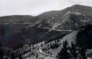A black and white photo of the railway going on the left side of a series of mountain peaks. Surrounding the trail are forests of pine trees.