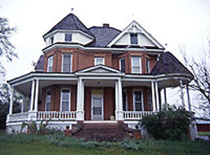 A view of a red bricked house with wrap-around porch, gabled entrance and roof, and a large octagonal turret and bay window on right. 