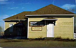 A view of the building with shaded single-door entrance and white trim. The building's shadow extends over its left side. 