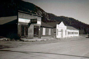 A black and white photo of a row of buildings on the side of a street in front of a series of hills.