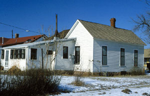 A view of the house from a corner. The yard in front is covered with snow and there is some leafless brush on the left. 