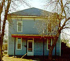 A front view of the house with hipped roof and covered porch. In front of the house stand two leafless trees. 