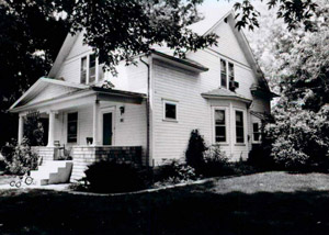 A black and white photo of the house with large gabled windows and porch. There is plenty of foliage around the photo. 