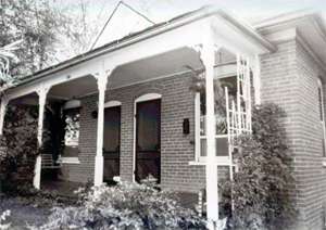 A black and white view of the brick building from a low angle, with white pillars holding up a covered porch in front.