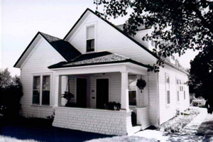 a black and white photo of the house with gabled roof and covered hip-roofed porch. 