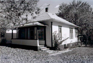 A black and white view of the house from an angle with hipped roof and covered porch with a path leading to it wrapping around the house. 