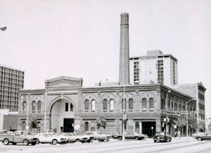 A black and white photo of the building with a large arch in the front, smokestack in the rear and cars parked in front. 