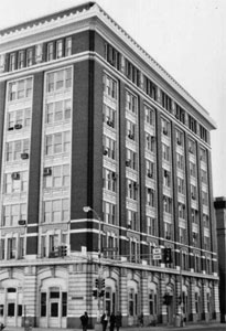 A black and white photo of the building with dark brick, white trim, large windows above and a light first floor. 