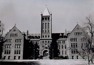 A view of the building with tall tower in the center and wings on either side with triangular roofs in black and white. 