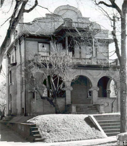 A black and white photo of the house with arch covered porch and stylized roof. There are leafless trees on both sides of photo and foreground and snow covering the yard. 