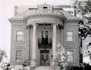 A black and white photo of the symmetrical building with tall pillars holding up a round balcony with pyramidal roof. On either side of the entrance are two layers of square windows and a flag in the center. 
