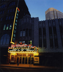 A view of the theater at night lit with various colors near the entrance and a vertical sign that gives the name of the building above the marquee. 
