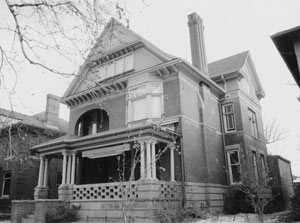 A black and white photo of the gabled house with windows in the garret, on the second floor is a bay window on the right with an arched balcony on the left and a large covered porch on the ground below with the entrance on the left. 