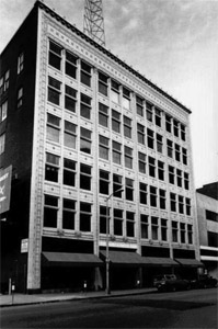 A black and white photo of the square building with ornate window facade and awning on street level. 