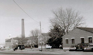 A black and white photo of a building on the left with a smokestack and a house on the right with a leafless tree and an evergreen in the center. 