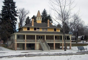 A view of a yellow pyramidal shaped building with large elevated wrap-around porch and stairs leading up to center. There are large evergreen and leafless trees also and snow-covered ground. 