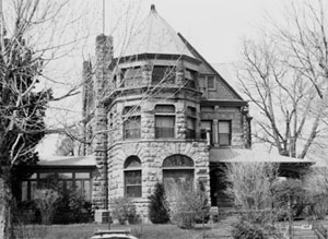 A black and white view of the house with an octagonal room in front with triangular roof overhead and windows on each wall. On either side are leafless trees and porch and the rest of the house on the right side. 