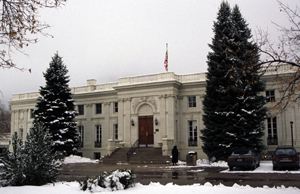 A view of the building with snow on the ground and two large pine trees on either side. The building stands in the back with tall windows and large entrance with staircase descending to a sidewalk below. 