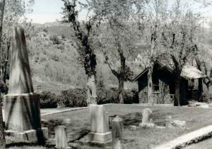 A blacka nd white photo of the cemetery with tombstones standing in the foreground and trees and foliage in the background. On the right stands a small building.