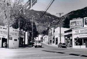 A black and white photo of the district with rows of commercial buildings on either side of a road, mountains i the background and power lines overhead. 