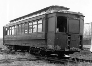 A black and white photo of the street car  sitting on tracks with a series of windows on the side and missing one on the front. 