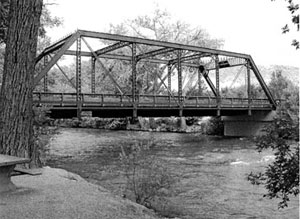 A black and white photo of the bridge with truss over the river. 