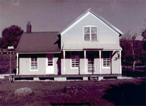 A direct view of the house with gabled roof, overhanging porch and slanted roof on the right. 