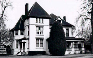 A black and white photo of the house with hipped roofs and white sides. A tall manicured push stands in the center. 