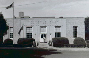 A black and white photo of the building with four bushes in front. 