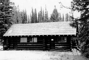 A black and white photo of the log walled house with pitched roof and tall pine trees in the background. Throughout the photo is a light layer of snow.