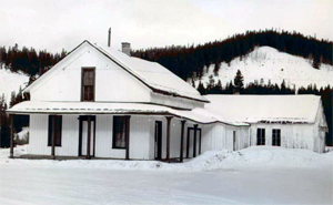 A black and white photo of the house with two story gable roof and covered porch on the left and cross roof on the right. There is a layer of snow on all surfaces and forested mountains in the background.
