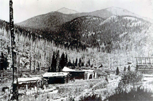 A black and white photo of the site with some buildings before snow covered evergreen trees and a mountain slightly in the distance.