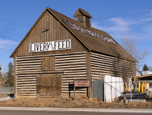 A picture of the barn with wood gabled roof that each have a sign that says "LIVERY AND FEED" painted on them. Above the barn is a small tower in the center with log walls below. 