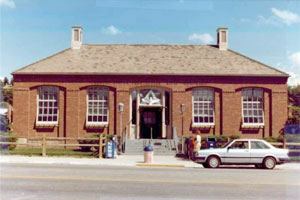 A picture of the building from the front with large windows, red walls and hipped roofs and the entrance in the center.