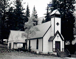 A photo of the church in black and white with steep gabled roof and cross room at the back. At the gabled entrance at the long end of the building is a small bell tower. 