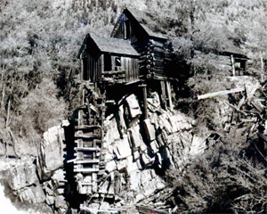 A photo of the mill with log sides, and pitched roof standing over a rock structure in black and white.