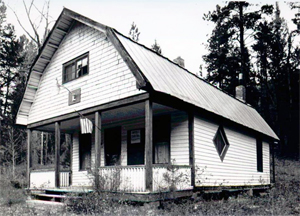 A photo of the building in black and white with gambrel roof overhanging a porch on the front and white siding. 