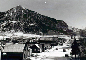 A black and white photo of the district with buildings and snow street on the bottom and right and a mountain peak on the left. 