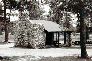 A black and white photo of a shelter made of stone with chimney and gabled roof, surrounded by trees.