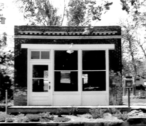 A black and white photo of the building with white trimmed door and windows below a flat roof.