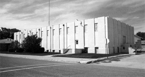 A black and white photo of the building with mostly bare facade and vertical depressions with windows on the top and bottom. 
