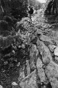 A black and white photo of a part of the trail with a sloping wall of boulders holding it up and a hiker standing on it. 