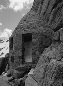 A black and white photo of the shelter with narrow entrance and stones leading up to it on the side of a mountain. 