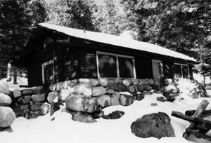 A black and white photo of the station with large stones at the base, gabled  roof, and wood siding. On the roof and around the station there is a layer of snow and large pine trees in the background.