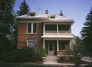 A photo of the house from the front with covered porch and covered balcony on the right and dormers on top. On either side stand very large evergreen trees and bush on the left of the entrance. 