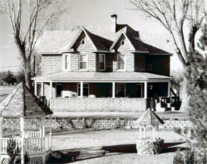 A photo of the house with twin cross gables above a long porch in black and white. In the foreground are a gazebo and a smaller well on the right both covered by octagonal cones between two leafless trees.