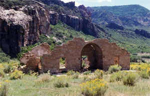 A picture of the mansion with some walls, a large archway and no roof in front of some rock formations and surrounded by grass.
