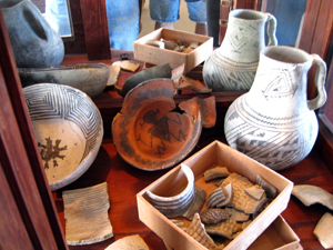 A collection of pottery inside a case with a pitcher on the left and bowls on the right before a box containing smaller fragments. There is a mirror in the back of the case reflecting everything within.